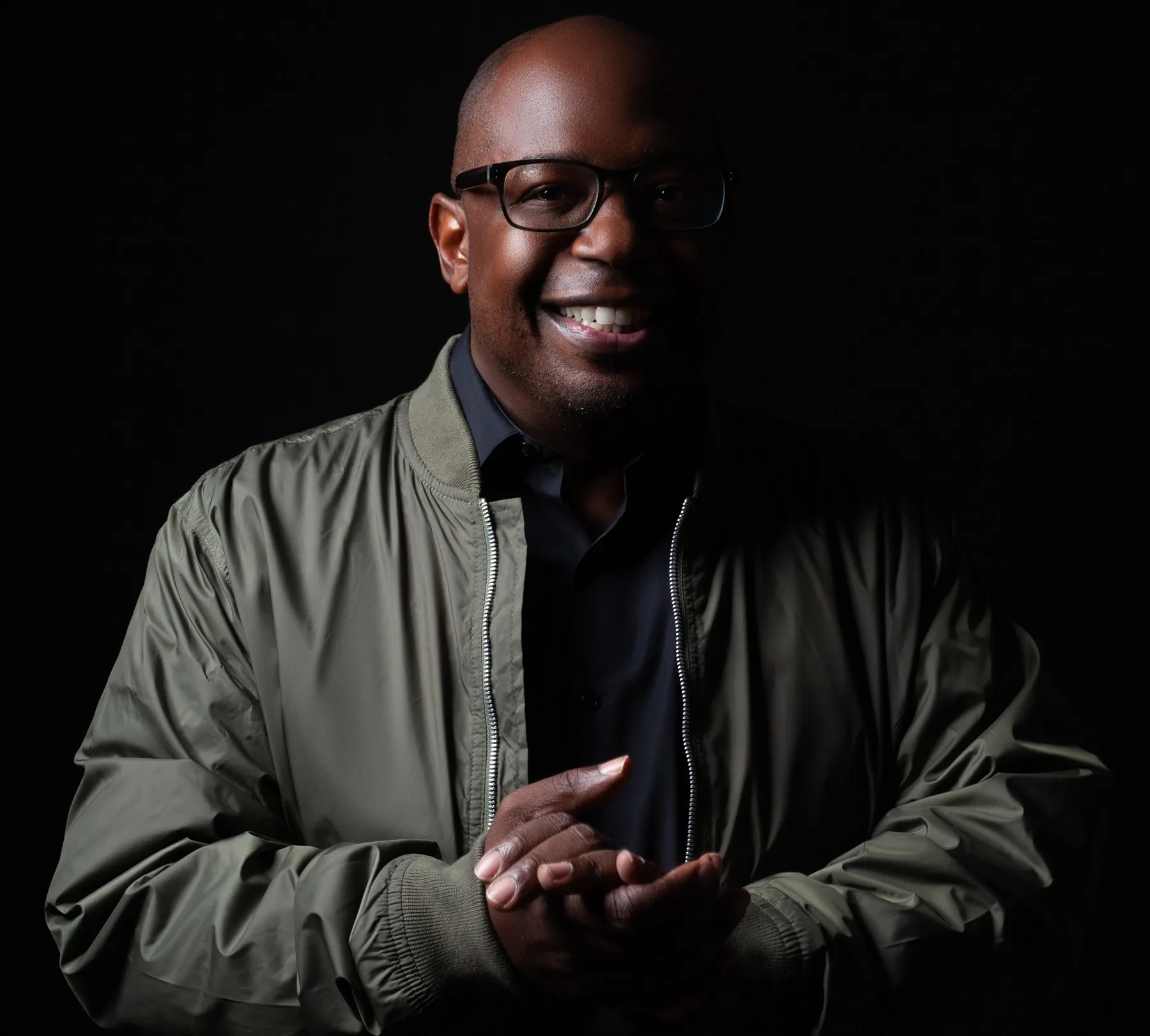 DJ Madd smiling in a green bomber jacket against a black background